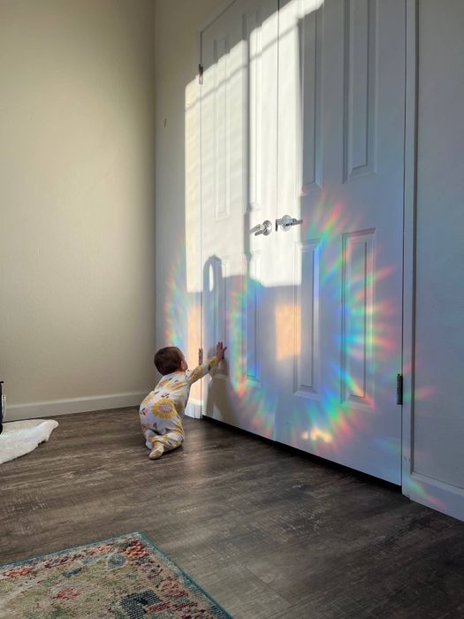 A toddler reaching out to touch the colorful light patterns on a door created by a prism, dressed in a patterned outfit