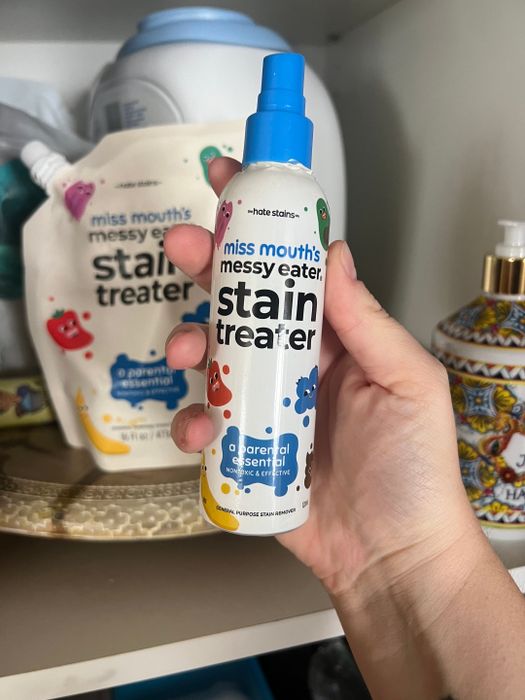 A hand holding a bottle of Miss Mouth's Messy Eater Stain Treater against a backdrop of various cleaning supplies