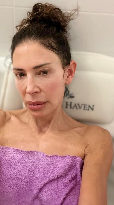 A person wrapped in a towel, taking a selfie in a bathtub with their hair up.