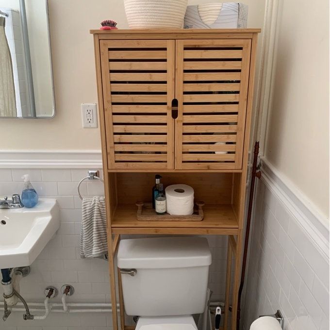 A reviewer's photo showing a wooden bathroom cabinet placed above the toilet, featuring closed upper doors and an open shelf displaying toiletries.
