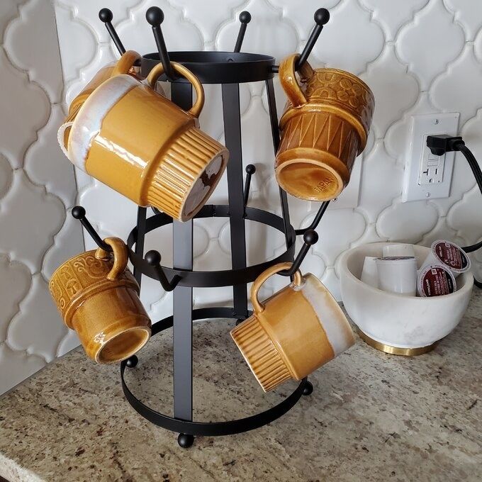 Four brown mugs rest on a metal mug tree on a kitchen counter, with a bowl of coffee pods beside them.