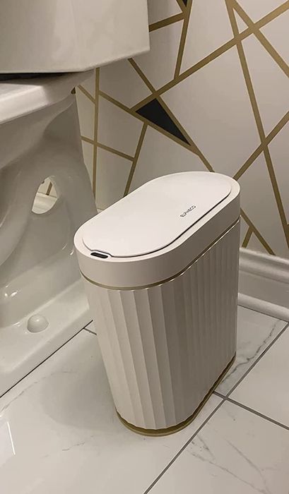 A white trash can with a sensor lid positioned next to a toilet in a bathroom