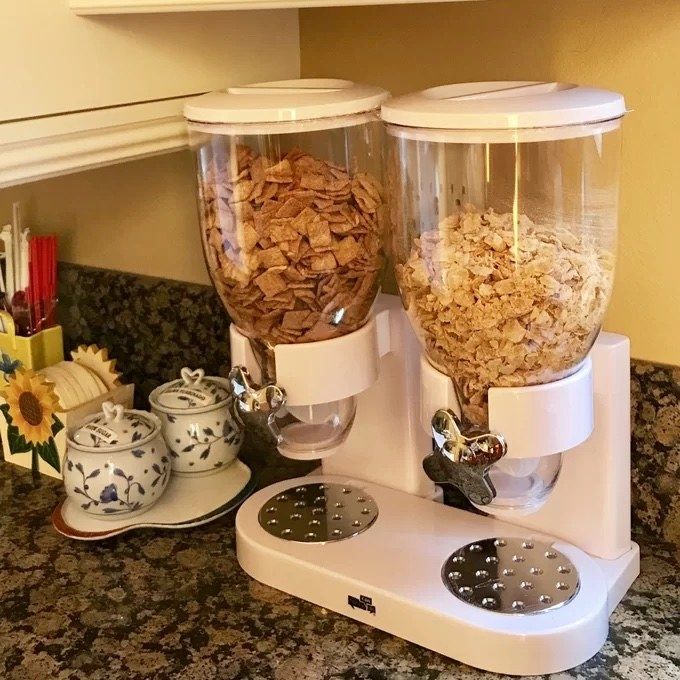 A kitchen countertop displays a dual cereal dispenser filled with cereals. Nearby are decorative ceramic jars and utensils.