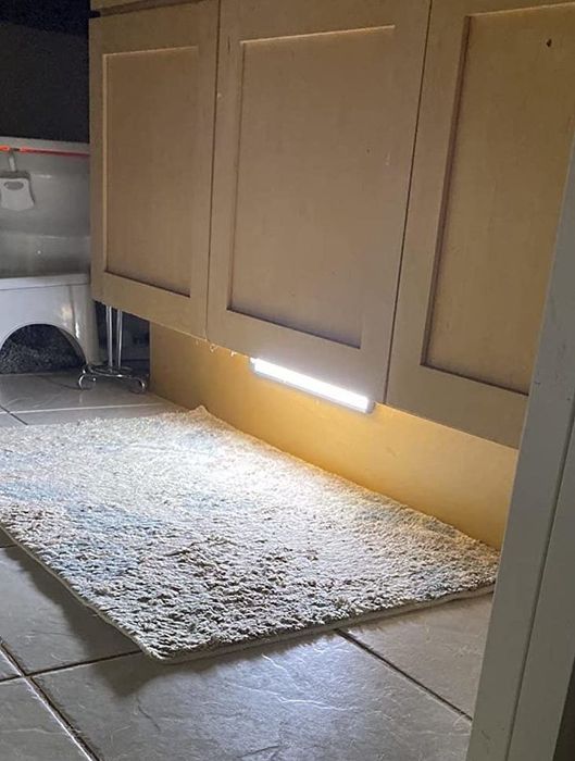 An under-cabinet LED light illuminating a kitchen mat on a tiled floor, with a partial view of cabinets and a container