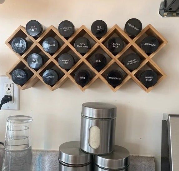 A wooden spice rack mounted on the wall, holding various labeled spice jars, positioned above a kitchen counter with three metallic canisters nearby.