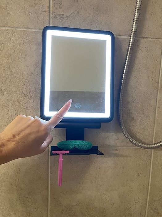 A finger presses a button on an illuminated bathroom mirror, with a razor and sponge neatly placed on a shelf below.
