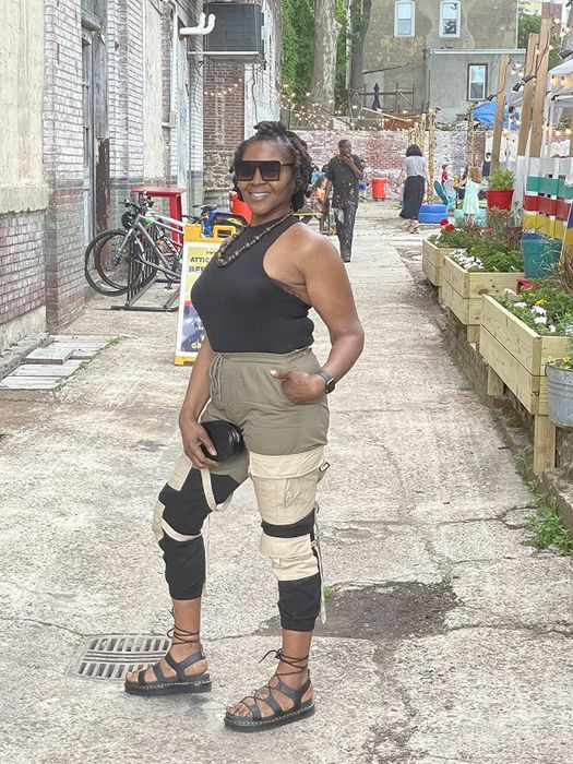The reviewer is dressed in a casual sleeveless top and cargo pants with knee patches, completing the look with a pair of strappy Dr. Martens sandals.