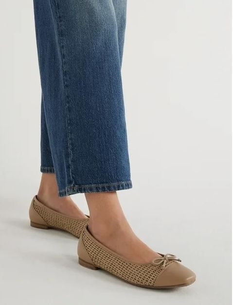 A woman wears classic beige perforated ballet flats paired with blue denim jeans.