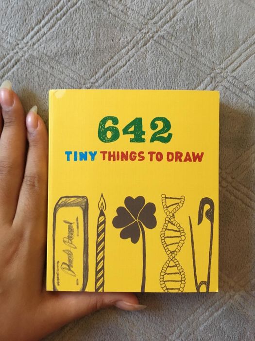 a compact book titled '642 Tiny Things to Draw' held delicately between fingers to emphasize its petite size