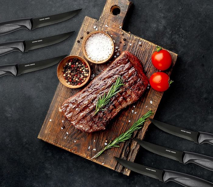 A cooked steak placed on a wooden cutting board surrounded by six steak knives
