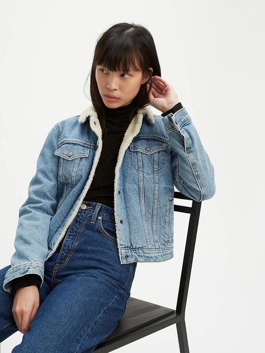 A model sitting on a chair wearing a light wash denim jacket with sherpa lining.
