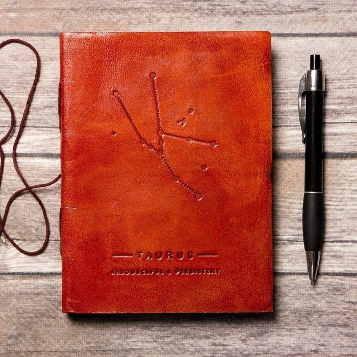 The Taurus brown leather journal, engraved with the adjectives resourceful and persistent, placed next to a pen