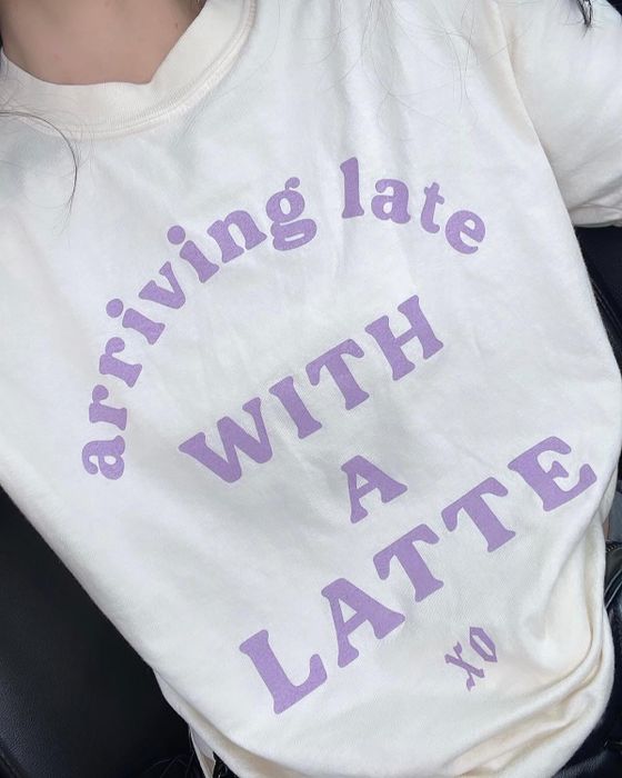 A tan graphic t-shirt featuring lavender text that reads, 