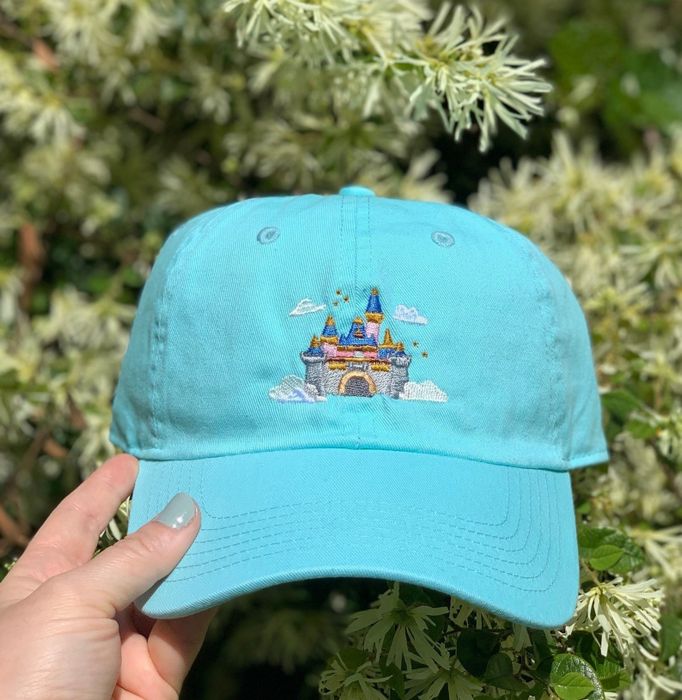 A teal hat adorned with an embroidery of Sleeping Beauty Castle floating among the clouds.