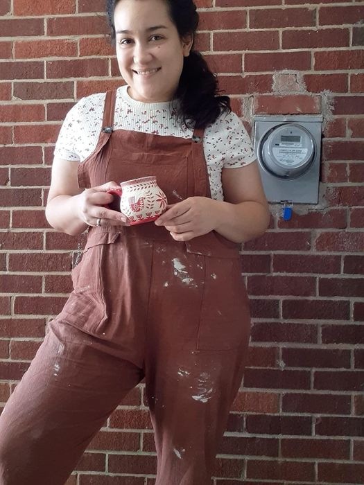 A reviewer wears overalls covered in clay splatters, layered over a T-shirt, and holds a small ceramic cup.