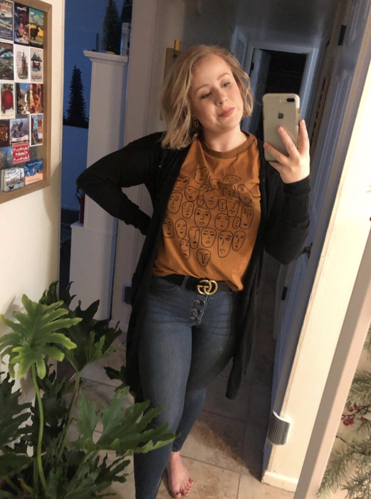 A reviewer models a dark yellow graphic tee featuring stylized line drawings of faces, paired with a black cardigan