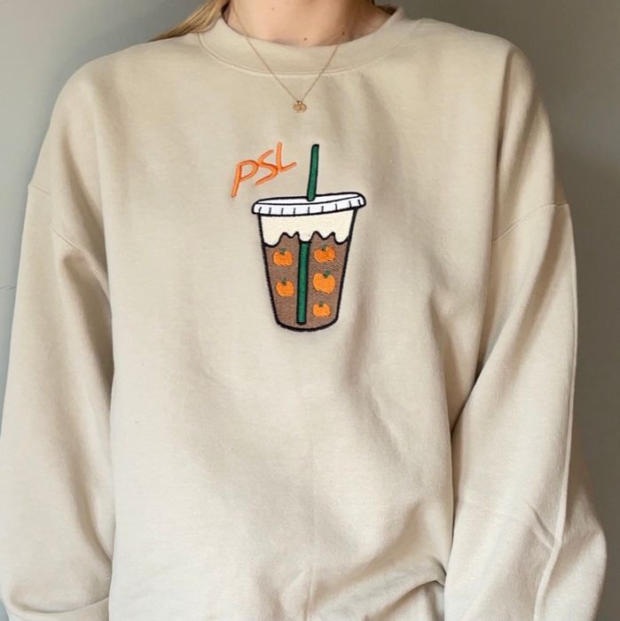 A tan sweater featuring an embroidered pumpkin spice latte with pumpkins floating inside it