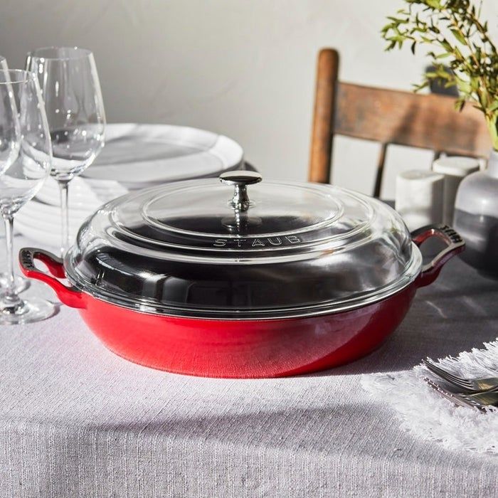 A Staub cast iron braiser with a glass lid, elegantly placed on a table alongside plates, glasses, and a decorative plant, showcasing its versatility for kitchenware enthusiasts