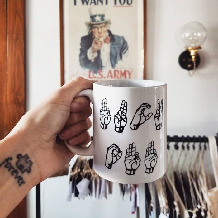 A white mug featuring an illustration of hands using American Sign Language to spell out the phrase 'Fuck Off'.