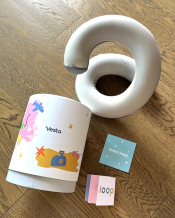 A neck pillow with Vesta branding, its packaging, and booklets resting on a wooden surface.