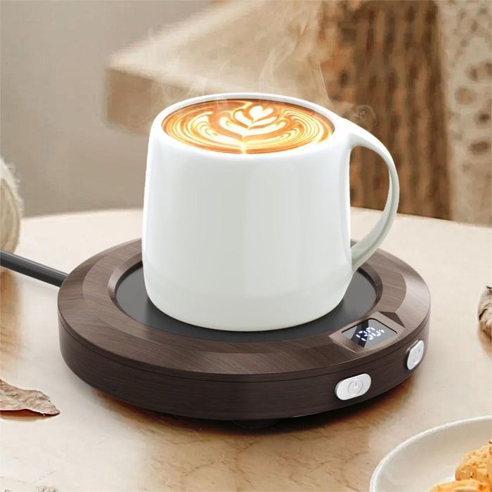 A hot latte with beautiful latte art rests on an electric mug warmer with a digital display, sitting on a wooden surface