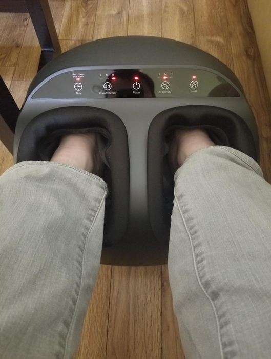 A close-up shot of feet inside a foot massager on a wooden floor, with visible control buttons for adjusting strength, power, air intensity, and heat settings.