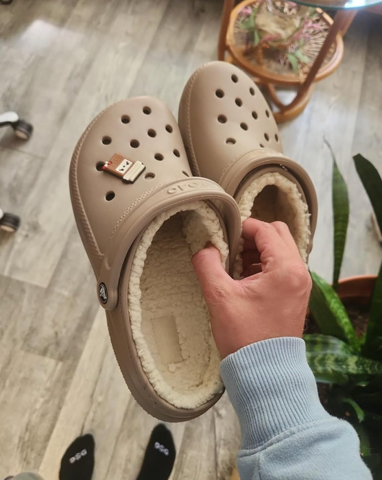 A hand holding a pair of beige Crocs adorned with a USB charm.