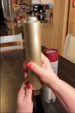 Hands twisting open a gold cylindrical canister with a twist-off cap, set on a table next to another container, within a cozy home environment.