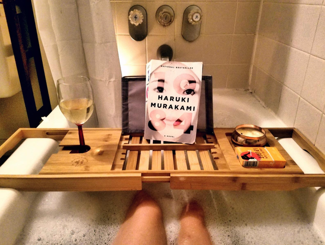 A person relaxes in a bathtub, with a bamboo tray across the edge, holding a glass of crisp white wine and a book titled