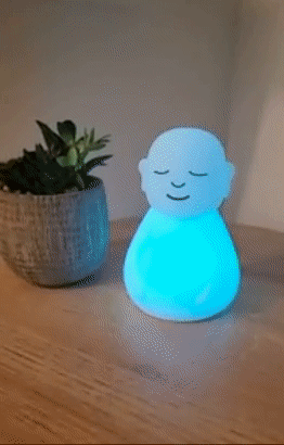 A small white figurine with closed eyes, its light shifting through shades of blue and green.