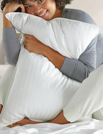 A person embraces a soft pillow, dressed in a cozy sweater and loose pants, smiling gently in a comfortable, inviting atmosphere.