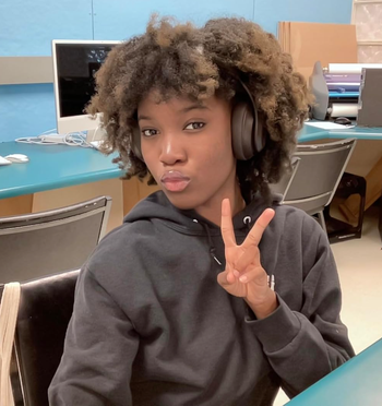 A person in a hoodie, wearing headphones, sitting at a desk and giving a peace sign, captured in a computer lab environment.
