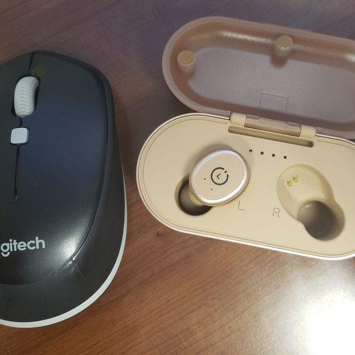 One of the rose gold wireless earbuds in its charging case, placed next to a Logitech mouse for size comparison