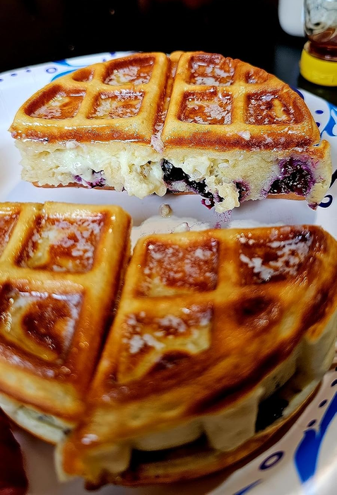 Close-up of waffles sliced open to reveal blueberries inside, served on a plate with maple syrup and strawberries in the background.