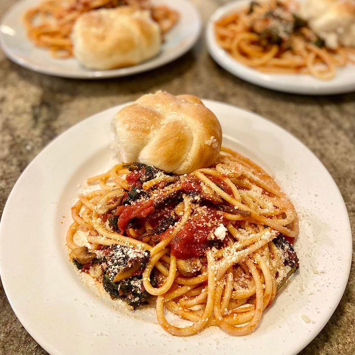 A plate of spaghetti topped with marinara sauce and freshly grated cheese, paired with a roll, placed on a table with other similar dishes in the background.