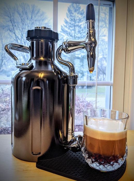 A stainless steel cold brew maker with a curved spout beside a freshly brewed cup of coffee
