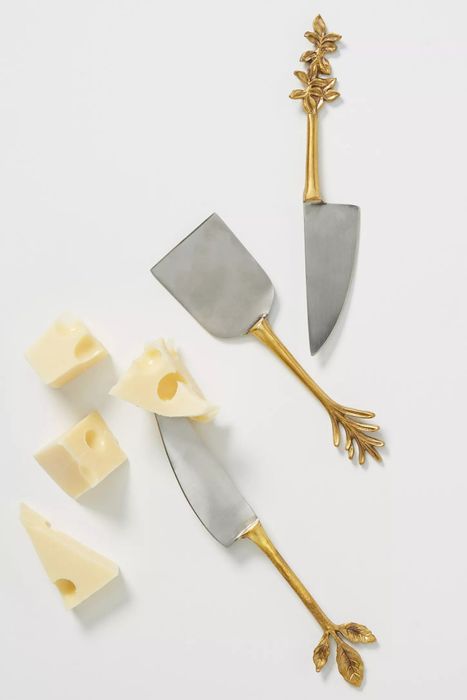 Three elegantly crafted silver and gold cheese knives with plant-shaped handles, positioned beside cubes of cheese.
