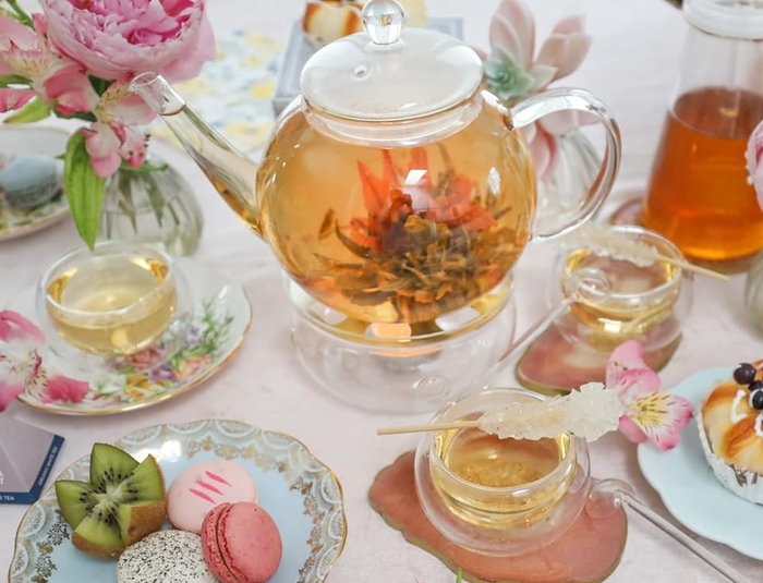 A sophisticated tea set featuring a glass teapot, floral-patterned cups, delectable pastries, and vibrant macarons, set on a beautifully decorated table, evoking a chic afternoon tea gathering.