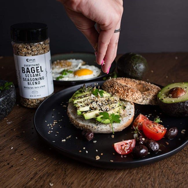 A jar of Everything Bagel Seasoning, with some of the seasoning being sprinkled onto a freshly toasted bagel.