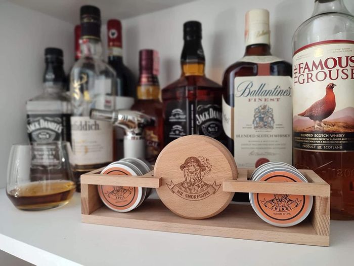 A display shelf showcasing an assortment of whiskey bottles paired with a set of wood chips designed for crafting smoked cocktails. A glass of whiskey rests beside the set.