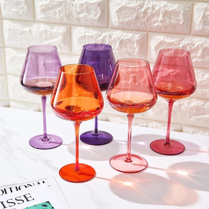 Five vibrant wine glasses placed on a marble countertop, accompanied by a nearby magazine. The glasses feature hues of purple, red, and orange.