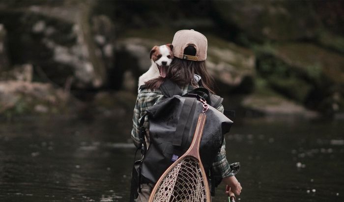 A person holding a dog in their arms, with the backpack strapped on their back, standing by a serene river.