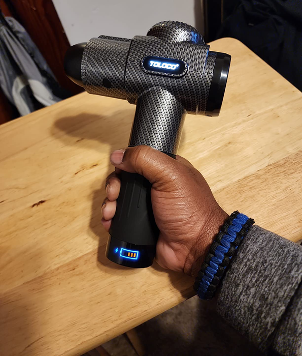The reviewer is shown holding a sleek, carbon gray massage gun.
