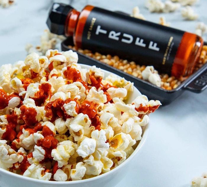 A bowl of popcorn drizzled with hot sauce, with the hot sauce bottle placed in the background.