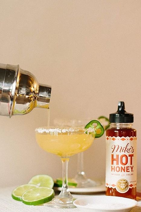 Someone pouring a cocktail from a shaker into a glass, with a bottle of Mike's Hot Honey placed beside the glass.