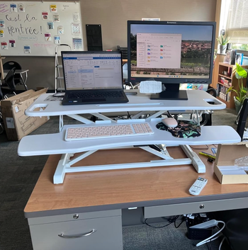 A white two-tier desk riser holding a keyboard on the lower shelf and a laptop and monitor above it on the higher shelf