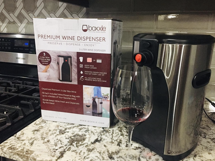 A reviewer photo showing the sleek silver wine dispenser on a counter, with a wine glass in front, pouring red wine into it.