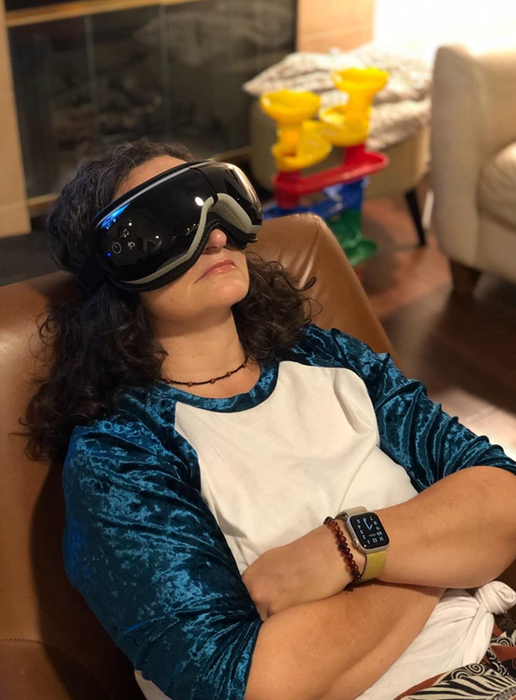 A reviewer wearing a black eye massager with gray cushioning around the nose, comfortably strapped to their head.