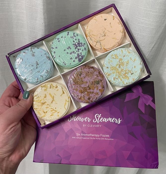 A reviewer holding a purple box filled with six vibrant shower steamers in various colors
