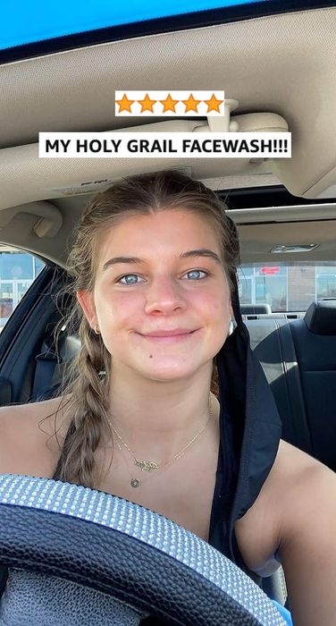 A reviewer with braided hair is sitting inside a car, wearing a necklace and a face mask that is partially pulled down. In the background, store signs are visible outside the vehicle.